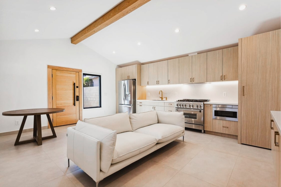 Bright and modern garage conversion ADU featuring a cozy living area with a sleek white couch, a spacious kitchen with light wood cabinets, stainless steel appliances, and a dining table, all illuminated by recessed lighting and accented with wooden beams.
