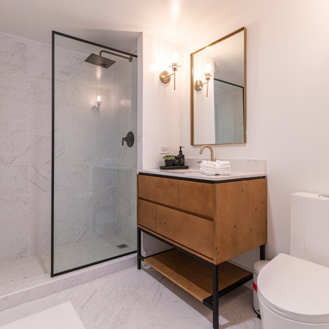 A luxurious bathroom in an ADU showcasing premium finishes. The space features a spacious glass-enclosed shower with elegant marble tile, a rain showerhead, and a sleek black frame. The wooden vanity with open shelving below is topped with a sophisticated countertop and gold fixtures, including a modern faucet. Above the vanity, a large rectangular mirror is flanked by stylish wall sconces that provide warm lighting. The bathroom exudes a blend of modern and rustic charm with its high-quality materials and meticulous design details.